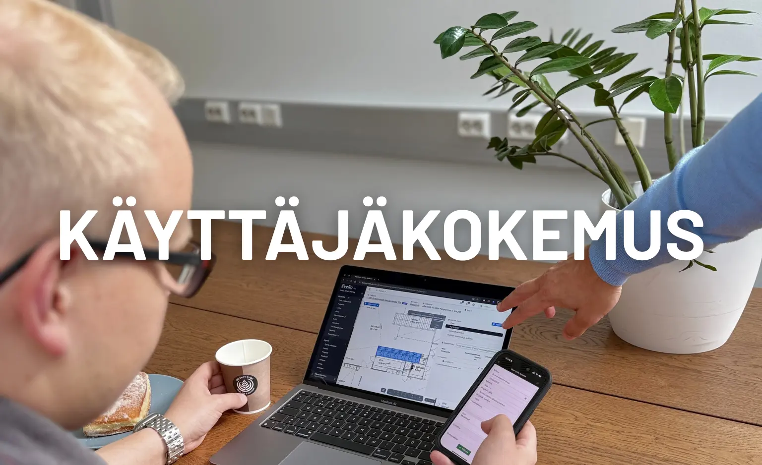 Kaksi henkilöä tarkastelee Evelian toiminnanohjausjärjestelmää läppärillä ja puhelimella. Toinen osoittaa näytöllä olevaa projektinäkymää, mikä korostaa ohjelmiston selkeää käyttäjäkokemusta.